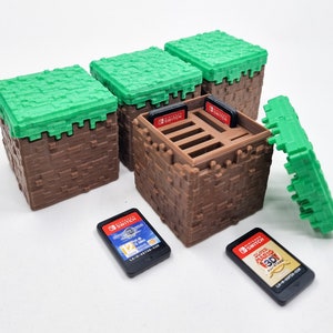May include: A set of brown and green pixelated storage cubes, resembling blocks. One cube is open, revealing slots for game cartridges. Two Nintendo Switch game cartridges are visible, along with a detached green top.