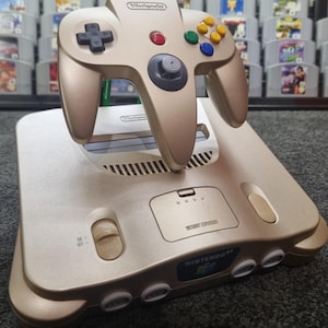 Könnte beinhalten: Eine goldfarbene Nintendo 64 Konsole mit passendem Controller. Der Controller hat ein Steuerkreuz, einen zentralen Analog-Stick und farbige Knöpfe. Die Konsole hat einen Power-Knopf und Steckplätze für Spielmodule. Eine Sammlung von Spielmodulen ist im Hintergrund zu sehen.