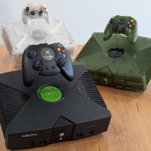 Peut inclure: Trois consoles Xbox avec leurs manettes assorties sont présentées sur une surface en bois. Les consoles sont noires, transparentes et vertes. La console noire a une manette noire, la console transparente a une manette transparente et la console verte a une manette verte.