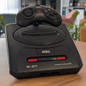 May include: A black Sega Genesis Model 2 video game console with a matching controller on top. The console has red and white accents and the text "16-BIT" and "MEGA DRIVE II".