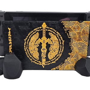 Könnte beinhalten: Schwarzer und goldener Videospielkonsolenständer mit einem Schwert- und Kreismuster, der eine schwarze Videospielkonsole hält. Der Ständer hat eine geometrische Basis und besteht aus einem strukturierten Material. Der Ständer ist für eine Spielekonsole konzipiert.