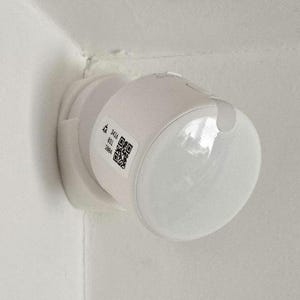 Op de afbeelding: Een witte, bewegingsgeactiveerde lamp die in een hoek is gemonteerd. De lamp heeft een afgeronde, heldere lens en een cilindrisch lichaam. Een sticker met tekst en een QR-code is zichtbaar.
