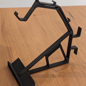 May include: Black plastic PS2 controller stand. The stand has a unique angled design with multiple support arms and a base. The text "PS2" is visible on the top arm. The stand is designed to hold a PS2 controller.