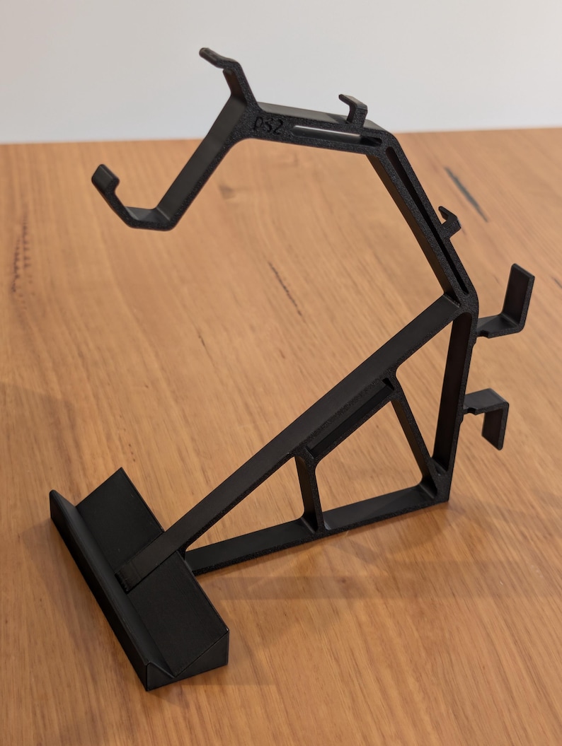 May include: Black plastic PS2 controller stand. The stand has a unique angled design with multiple support arms and a base. The text "PS2" is visible on the top arm. The stand is designed to hold a PS2 controller.