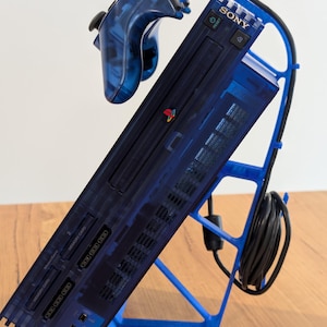 May include: A transparent blue PlayStation 2 console and controller are displayed on a blue stand. The console is angled, showcasing its ports and the Sony logo. The controller is connected, and the stand neatly organizes the console and its accessories.