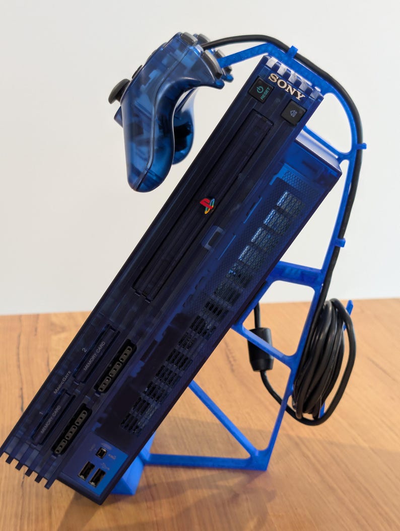 May include: A transparent blue PlayStation 2 console and controller are displayed on a blue stand. The console is angled, showcasing its ports and the Sony logo. The controller is connected, and the stand neatly organizes the console and its accessories.