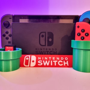 Puede incluir: Una consola Nintendo Switch con mandos Joy-Con rojos y azules, un cartucho de juego y un logotipo de Nintendo Switch rojo y blanco. Los mandos y el cartucho de juego se colocan en soportes verdes con forma de tubo.