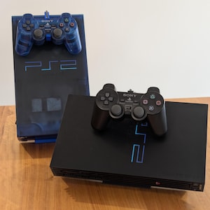 May include: Two PlayStation 2 consoles are displayed. One is black with a black controller, and the other is a translucent blue with a matching blue controller. The consoles are on a wooden surface. The text "PS2" is visible on both consoles.