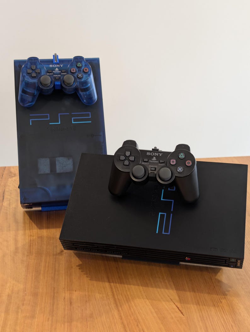 May include: Two PlayStation 2 consoles are displayed. One is black with a black controller, and the other is a translucent blue with a matching blue controller. The consoles are on a wooden surface. The text "PS2" is visible on both consoles.
