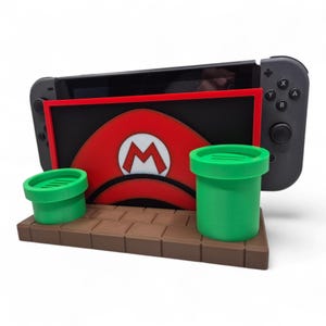 Puede incluir: Un soporte para consola Nintendo Switch con un sombrero rojo de Mario y dos tuberías verdes. El soporte marrón mantiene la consola en posición vertical. El sombrero tiene una "M" blanca.