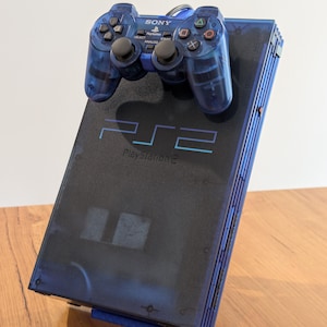 May include: A blue PlayStation 2 console with a matching blue controller. The console has the text "PS2 PlayStation 2" printed on the top. The controller has the Sony logo and various buttons.