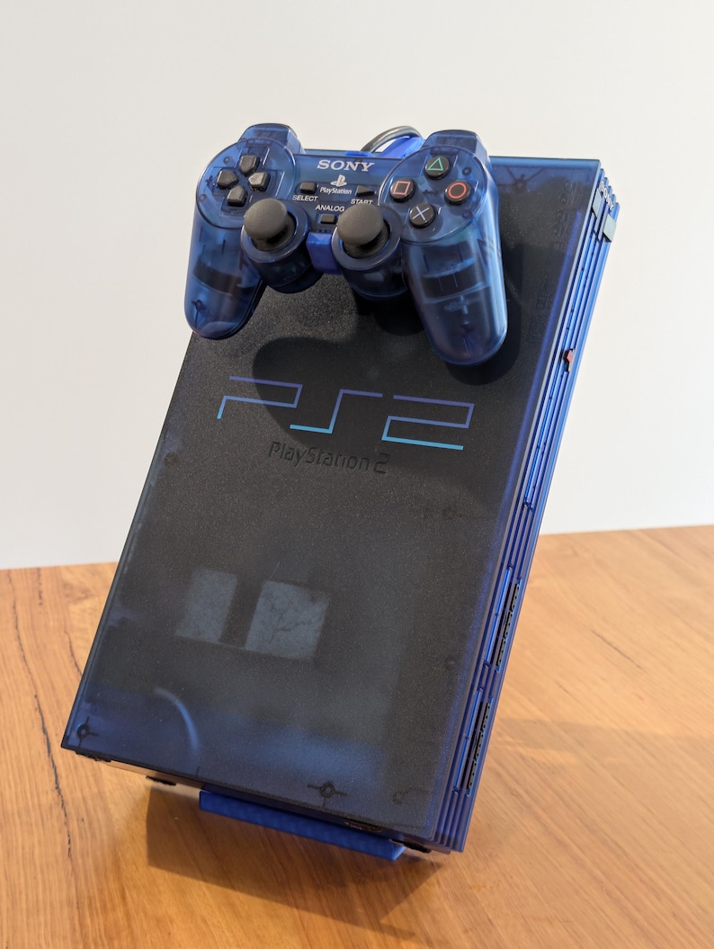 May include: A blue PlayStation 2 console with a matching blue controller. The console has the text "PS2 PlayStation 2" printed on the top. The controller has the Sony logo and various buttons.