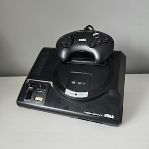 Puede incluir: Una consola Sega Mega Drive negra con un mando a juego. La consola tiene el texto "16-BIT" y "MEGA DRIVE SEGA" impreso. El mando tiene el logotipo de Sega y varios botones.
