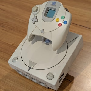 Puede incluir: Una consola de videojuegos Sega Dreamcast blanca con un mando a juego. El mando tiene una cruceta, una pequeña pantalla y botones de acción de colores. La consola tiene una bandeja de disco y varios puertos.