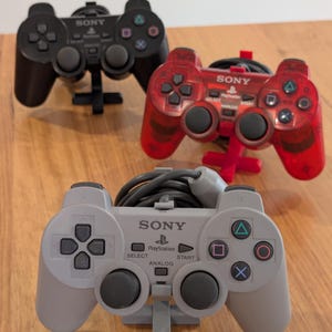 Peut inclure: Trois manettes Sony PlayStation noires, rouges et grises. Les manettes comportent des boutons directionnels, des boutons d'action et des joysticks analogiques. La manette grise porte l'inscription "SONY PlayStation" et "SELECT ANALOG START".
