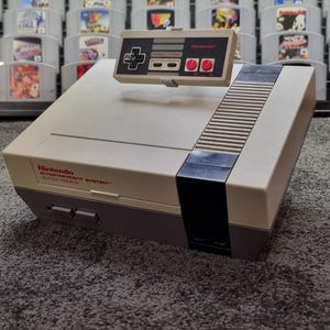 May include: A vintage Nintendo Entertainment System console with a matching controller. The console is beige and gray, with the Nintendo logo and text on the front. The controller is gray with black buttons and a directional pad. A collection of game cartridges is in the background.