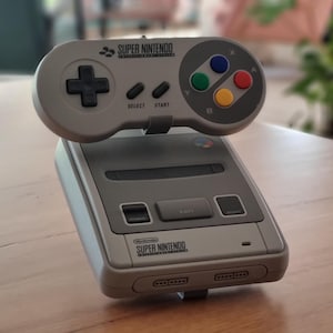 Könnte beinhalten: Eine graue Super Nintendo Konsole und Controller. Der Controller hat ein Steuerkreuz, Select- und Start-Tasten sowie farbige Tasten. Konsole und Controller befinden sich auf einer Holzoberfläche.