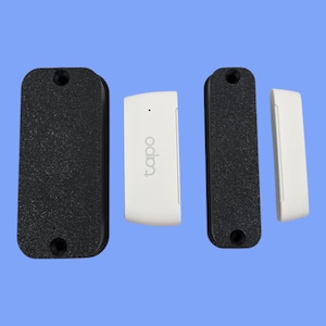 May include: A smart home security sensor set, comprising two black textured rectangular pieces with mounting holes and two white rectangular pieces. The white pieces have the word "tapo" printed on them.