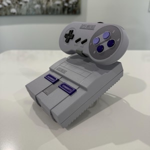 May include: A grey Super Nintendo console and controller. The controller has a directional pad, buttons, and the text "SUPER NINTENDO". The console has purple buttons and the text "SUPER NINTENDO".