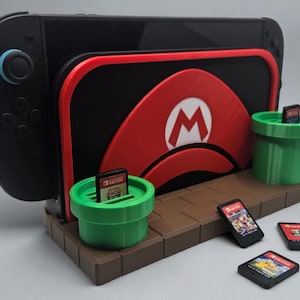 Op de afbeelding: Een zwarte Nintendo Switch-console staat in een aangepaste standaard met een rood en zwart Mario-thema-ontwerp. Twee groene, pijpvormige spelcassettehouders en verschillende spelcassettes zijn ook zichtbaar. De standaard is bruin.