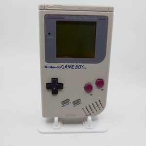 Puede incluir: Una consola portátil Nintendo Game Boy de color gris claro. El dispositivo presenta un mando direccional negro, botones A y B rosas y una pantalla que muestra una imagen oscura. El texto "Nintendo GAME BOY" es visible.
