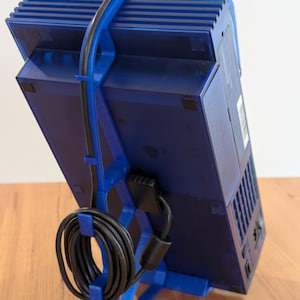 Haotogel Login May include: A blue gaming console is held by a blue 3D-printed stand Link Alternatif. A black power cord is attached to the console and is looped around the stand. The console is angled, showcasing its design and features.