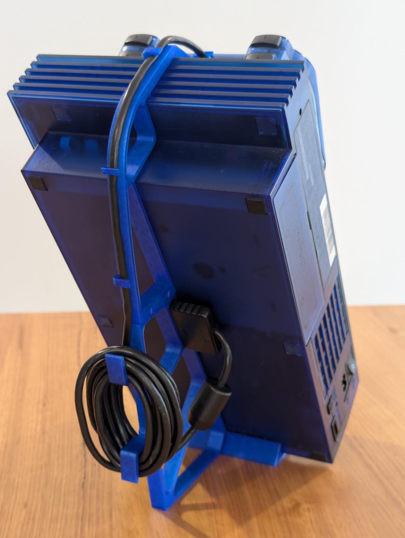 Haotogel Login May include: A blue gaming console is held by a blue 3D-printed stand Link Alternatif. A black power cord is attached to the console and is looped around the stand. The console is angled, showcasing its design and features.