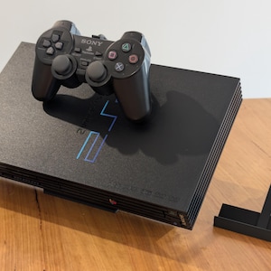 May include: A black PlayStation 2 console with a matching controller and a black stand. The console has a silver logo and the controller has the Sony logo. The console and controller are on a wooden surface.