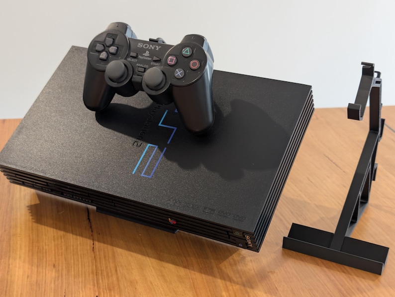May include: A black PlayStation 2 console with a matching controller and a black stand. The console has a silver logo and the controller has the Sony logo. The console and controller are on a wooden surface.