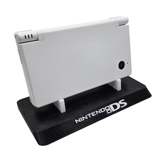 Op de afbeelding: Een witte Nintendo DS handheld gameconsole wordt weergegeven op een zwarte standaard. De standaard heeft de woorden "NINTENDO DS" in het wit gedrukt. De console staat in gesloten positie.