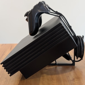 May include: Black video game console with a matching controller resting on top. The console is angled, revealing its layered design. A black stand supports the console, and a black cable is coiled nearby. The background is a light color.
