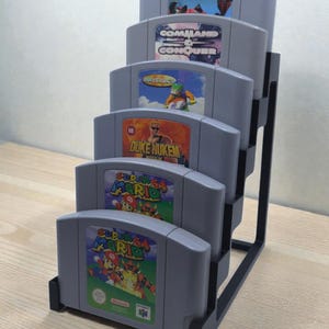 Peut inclure: Un présentoir en métal noir contenant six cartouches de jeu Nintendo 64 grises. Les cartouches présentent des titres de jeux tels que "Super Mario 64", "Duke Nukem" et "Command & Conquer". Le présentoir est sur une surface en bois clair.