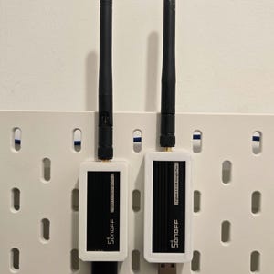 Può includere: Due dispositivi Sonoff Zigbee 3.0 USB Dongle Plus bianchi e neri con antenne nere. Uno ha un cavo USB, l'altro un connettore USB. I dispositivi sono montati su una pannello forato bianco.