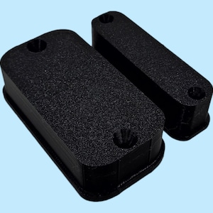 May include: Two black rectangular objects with rounded edges and textured surfaces. Each has a hole on the top surface. One object is larger than the other. The objects are on a light blue background.