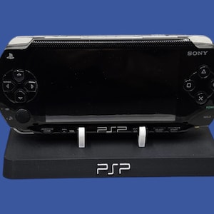 Może przedstawiać: Czarna konsola do gier przenośna Sony PSP spoczywająca na czarnym stojaku. Konsola ma błyszczące wykończenie z przyciskami sterowania i ekranem. Na stojaku widnieje napis "PSP".