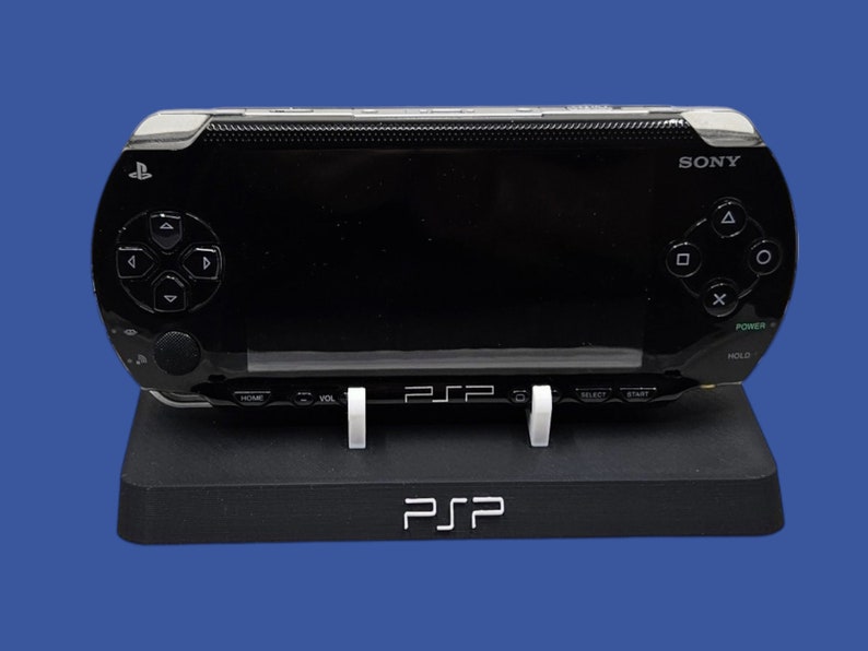 Pu&ograve; includere: Una console di gioco portatile Sony PSP nera appoggiata su un supporto nero. La console ha una finitura lucida con pulsanti di controllo e uno schermo. Il supporto ha le lettere "PSP" stampate.