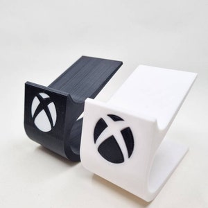 May include: Two 3D-printed phone stands, one black and one white. Each stand features a curved design with an Xbox logo. The stands are designed to hold a phone or other small device.