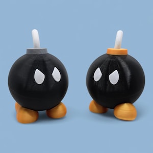 Puede incluir: Dos figuras esféricas negras que se asemejan a bombas de dibujos animados. Cada una tiene detalles blancos en forma de ojo, pies naranjas y una mecha blanca. Una tiene la parte superior gris, la otra naranja. El fondo es azul claro liso.