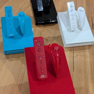 Puede incluir: Consolas Wii y mandos en varios colores: rojo, azul, negro y blanco. Cada consola tiene un mando y un nunchuk a juego. Los mandos tienen botones y una cruceta direccional. Sobre una superficie de madera.