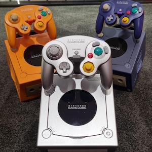 Peut inclure: Trois consoles et manettes Nintendo GameCube de couleur orange, argent et violet. Les manettes ont des boutons et des joysticks colorés. Les consoles sont présentées sur une surface sombre avec un arrière-plan d'étuis de jeux vidéo.
