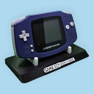 May include: A blue Game Boy Advance handheld video game console on a black stand. The console has a screen, buttons, and the text "GAME BOY ADVANCE". The stand also has the text "GAME BOY ADVANCE".