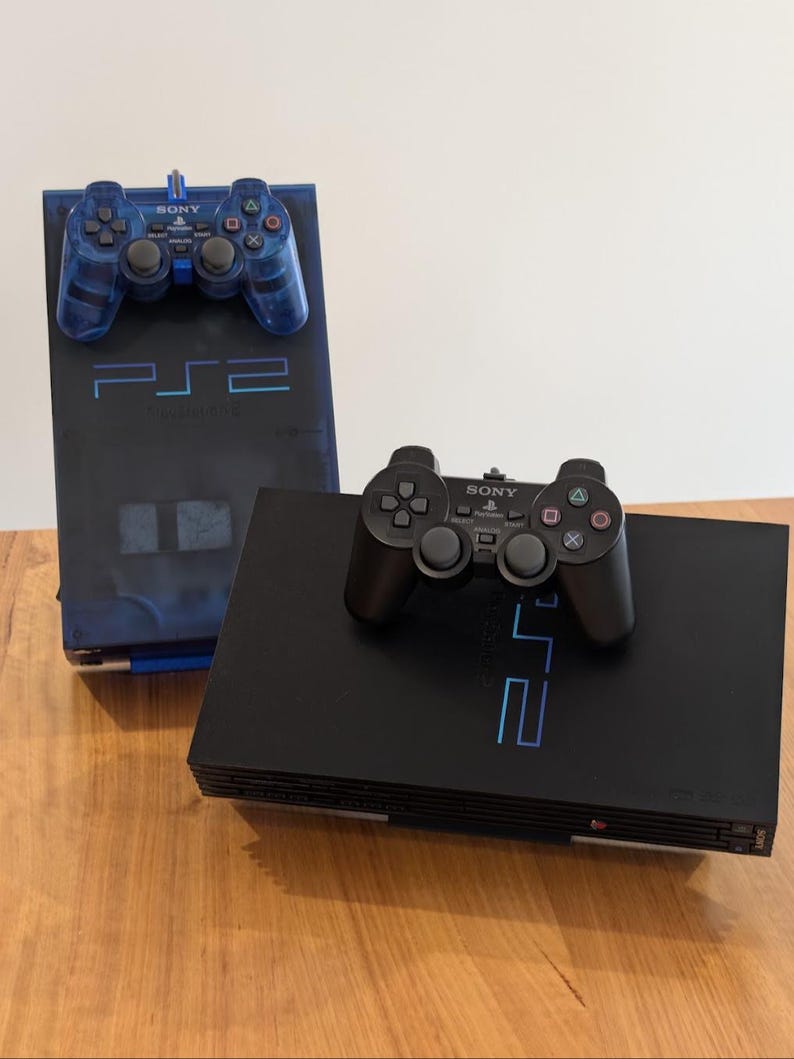 May include: Two PlayStation 2 consoles are displayed, one black and one blue. The black console has a black controller on top. The blue console has a blue controller on top. Both consoles have the PS2 logo.