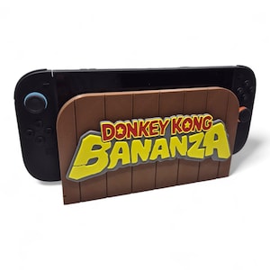 May include: A black Nintendo Switch console is held by a brown wooden stand with the words "DONKEY KONG BANANZA" in yellow and red lettering. The stand has a wood-grain texture.