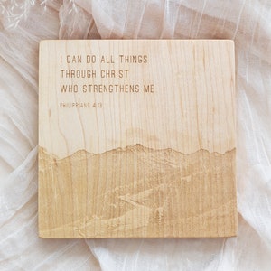 May include: A square wooden plaque with engraved text and a mountain range design. The text reads "I CAN DO ALL THINGS THROUGH CHRIST WHO STRENGTHENS ME" and "PHILIPPIANS 4:13". The plaque is light brown and sits on a white, textured surface.