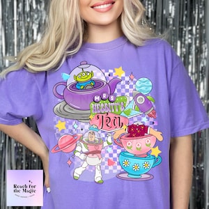 Puede incluir: Camiseta morada con un diseño gráfico que presenta una tetera espacial de dibujos animados, un astronauta de dibujos animados sosteniendo una taza de té y un letrero que dice "Mrs. Nesbitt's Tea".