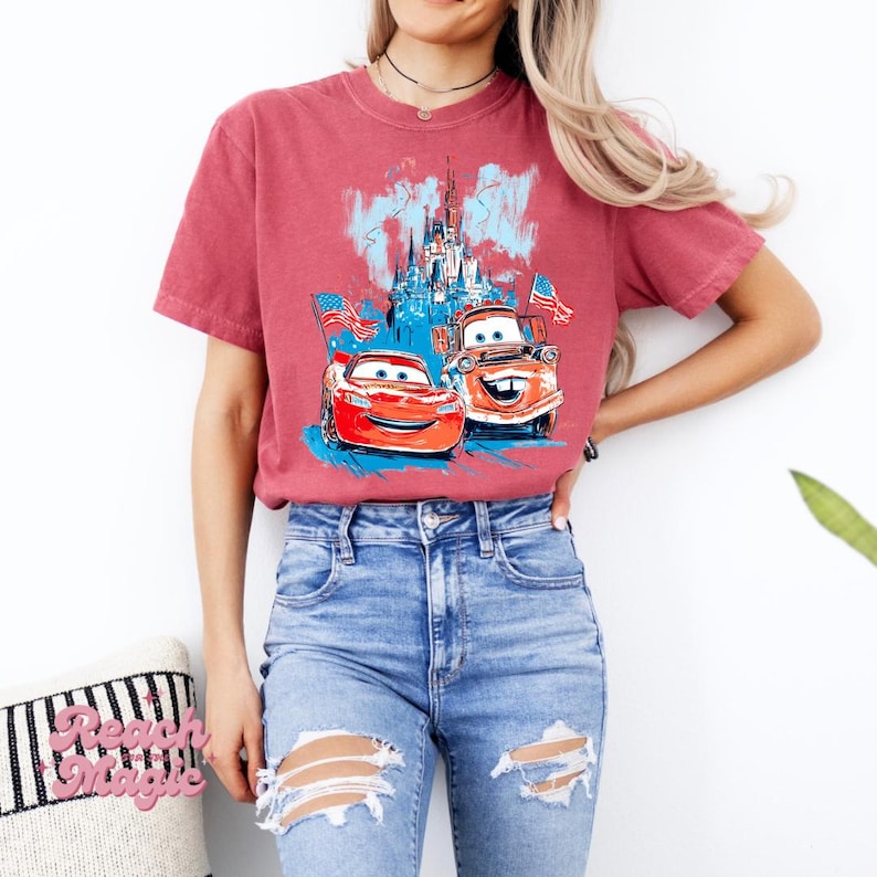 May include: Faded red t-shirt with a graphic of two cartoon cars with American flags in front of a castle. The shirt is paired with blue jeans.
