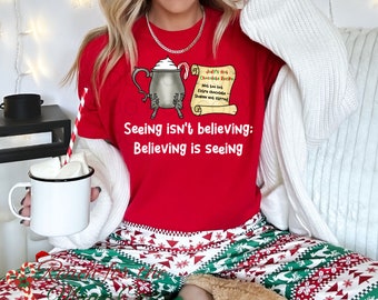 May include: Red t-shirt with a cartoon illustration of a hot chocolate pot and a scroll with the text "Judy's Hot Chocolate Recipe:  Milk, hot, extra chocolate, shaken not stirred." The shirt also has the text "Seeing isn't believing: Believing is seeing."