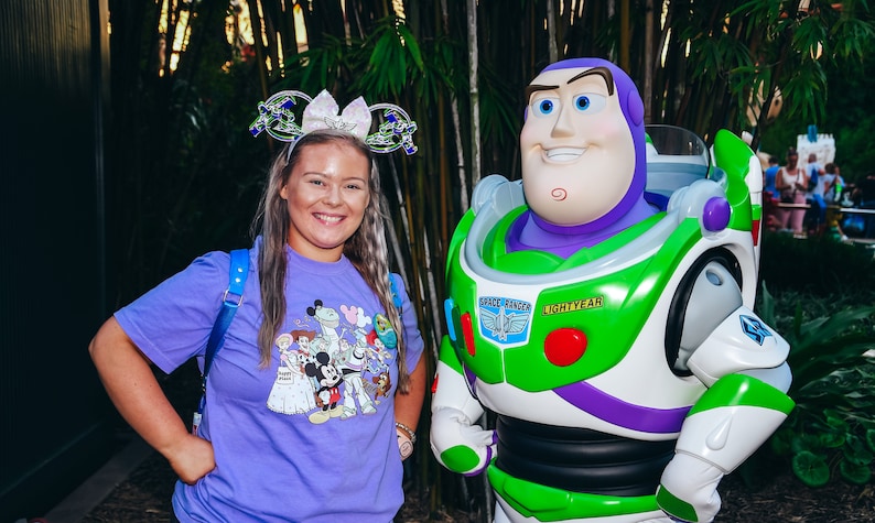 May include: A woman wearing a purple shirt with a cartoon character design stands next to a green, white and purple Buzz Lightyear character. The character has a large head and a friendly expression. The woman is wearing a sparkly headband with a crown on top.