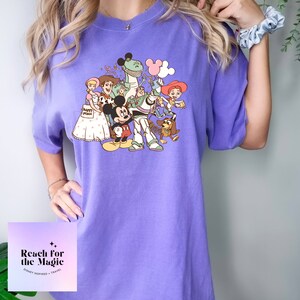 May include: Purple t-shirt featuring a vintage-style graphic of Mickey Mouse and the Toy Story characters, including Woody, Buzz Lightyear, Rex, and Slinky Dog. The text "Reach for the Magic" is printed on the bottom of the shirt.