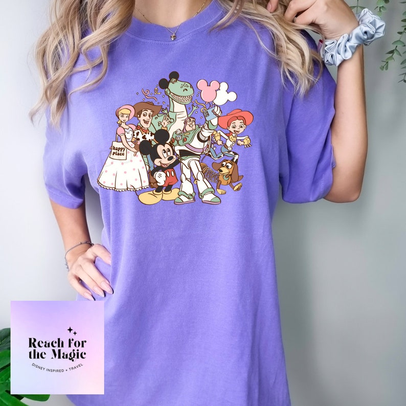 May include: Purple t-shirt featuring a vintage-style graphic of Mickey Mouse and the Toy Story characters, including Woody, Buzz Lightyear, Rex, and Slinky Dog. The text "Reach for the Magic" is printed on the bottom of the shirt.